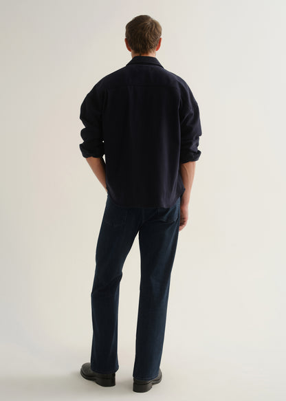 Person wearing a dark navy long sleeve button-up shirt with dark blue jeans and black boots, back full body view on a light grey background.