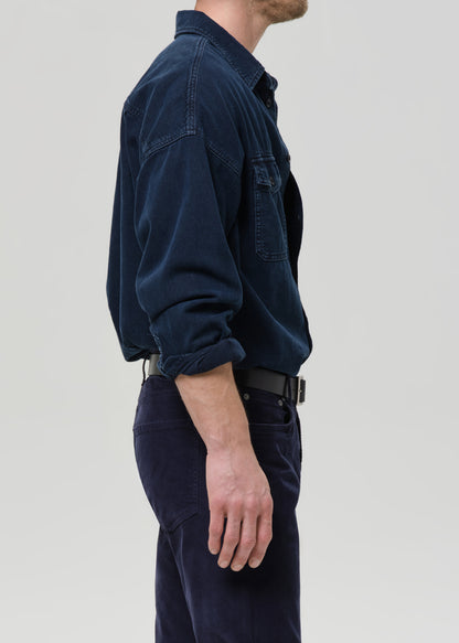 Man wearing a dark navy button-up shirt with dark navy straight leg pants viewed from the side on a grey background.
