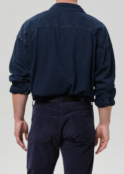 Man wearing a dark navy button-up shirt with dark navy straight leg pants viewed from the back on a grey background.