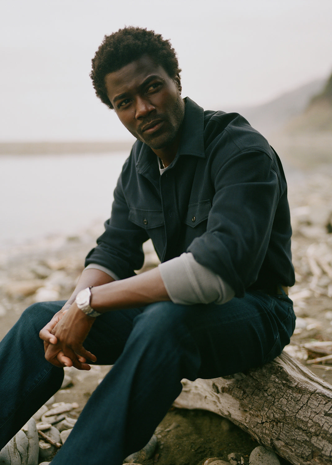 A man wearing a dark navy blue button-up shirt and matching navy jeans sits on a log on a rocky, beach-like shore with a soft-focus natural background.