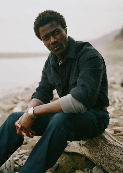 A man wearing a dark navy blue button-up shirt and matching navy jeans sits on a log on a rocky, beach-like shore with a soft-focus natural background.