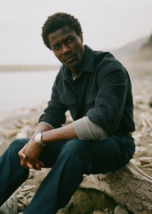 A man wearing a dark navy blue button-up shirt and matching navy jeans sits on a log on a rocky, beach-like shore with a soft-focus natural background.