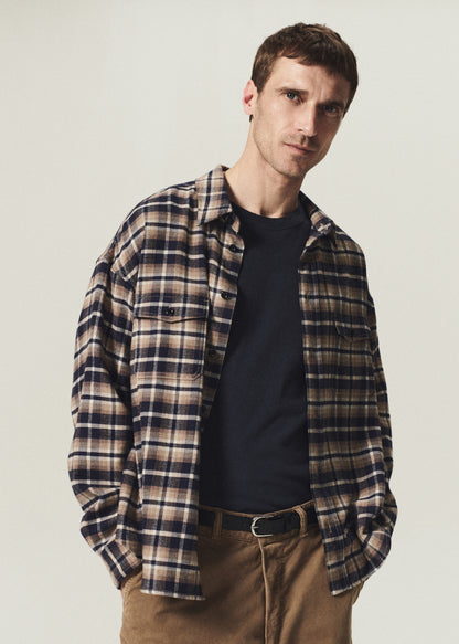 Man wearing a navy and brown plaid button-up shirt layered over a navy crewneck top with brown pants on a grey background.