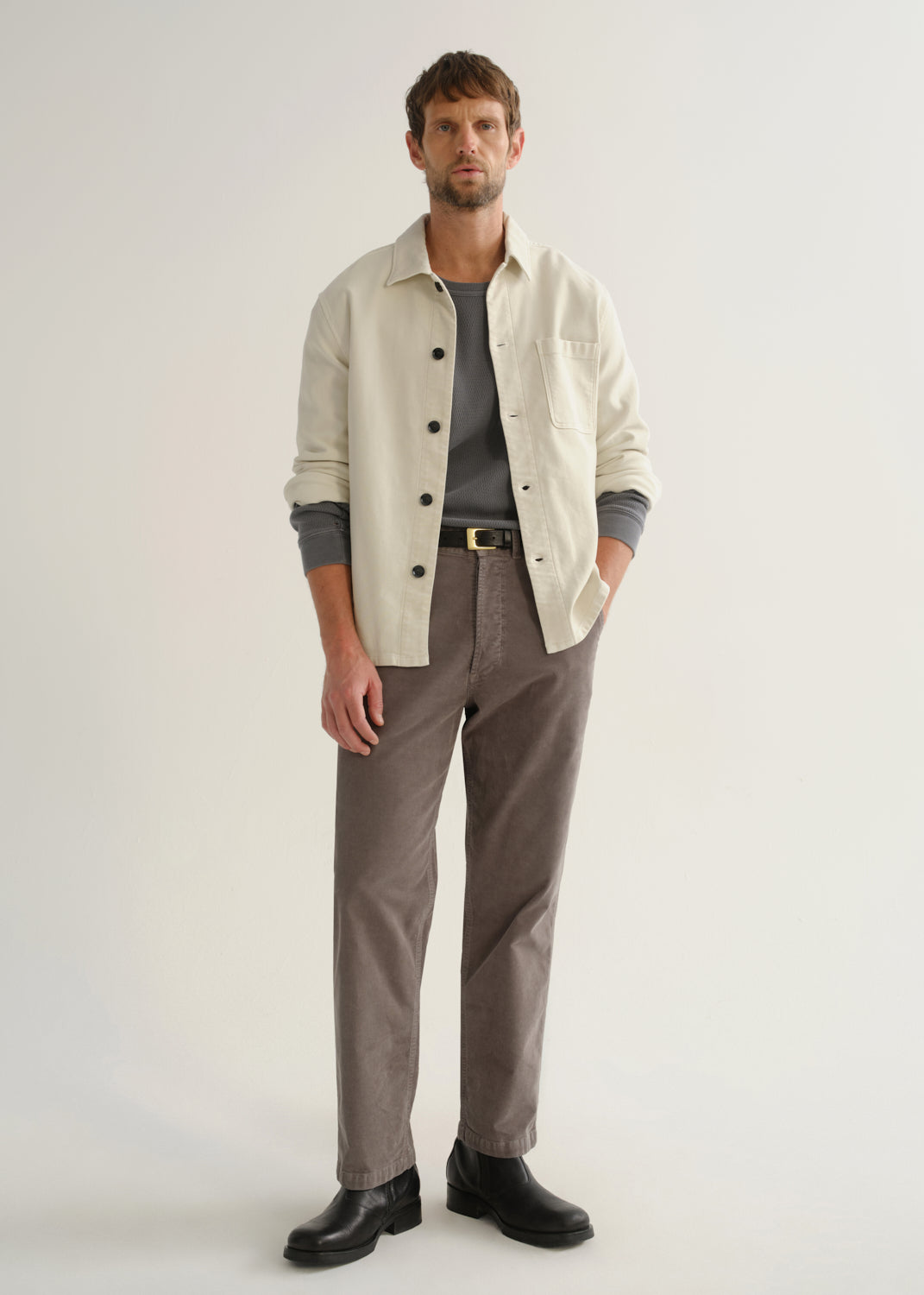 Person wearing a cream long sleeve button-up shirt over a grey top with brown pants and black boots, front full body view on a light grey background.