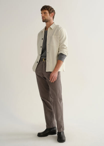 Person wearing a cream long sleeve button-up shirt over a grey top with brown pants and black boots, angled front full body view on a light grey background.