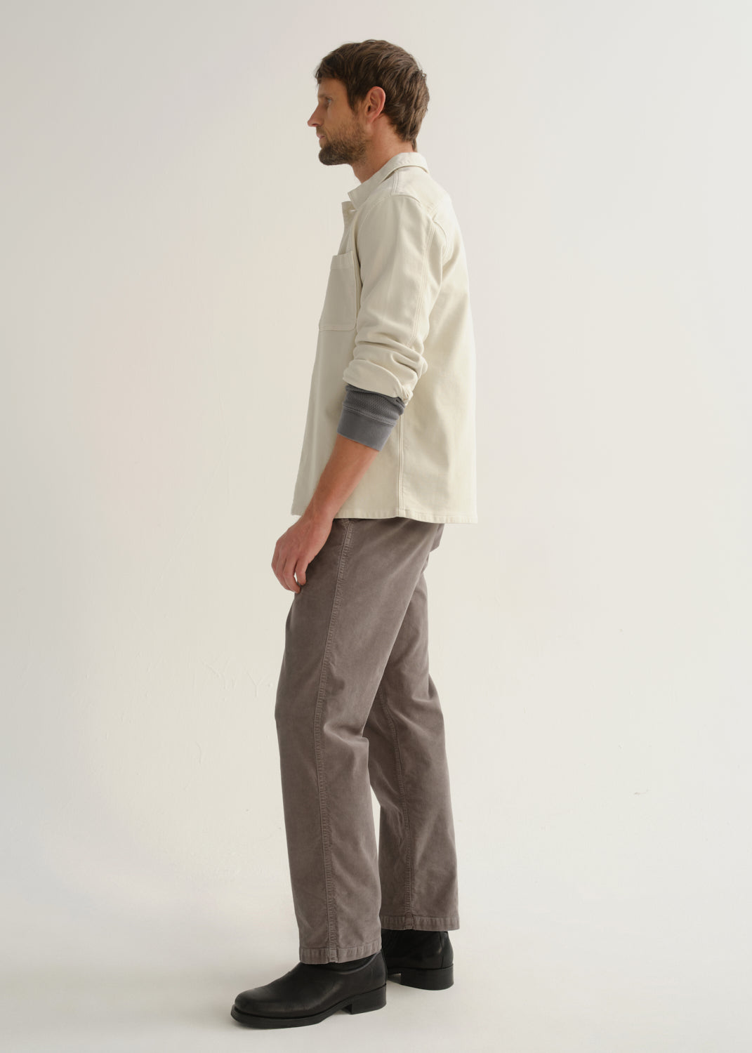 Person wearing a cream long sleeve button-up shirt with brown pants and black boots, side full body view on a light grey background.