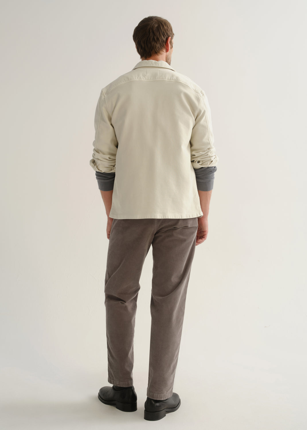 Person wearing a cream long sleeve button-up shirt with brown pants and black boots, back full body view on a light grey background.