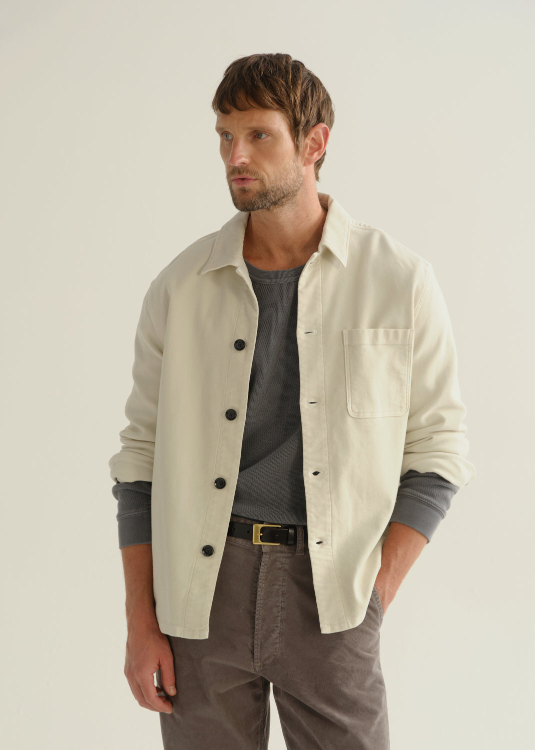 Person wearing a cream long sleeve button-up shirt over a grey top with brown pants, front cropped view on a light grey background.