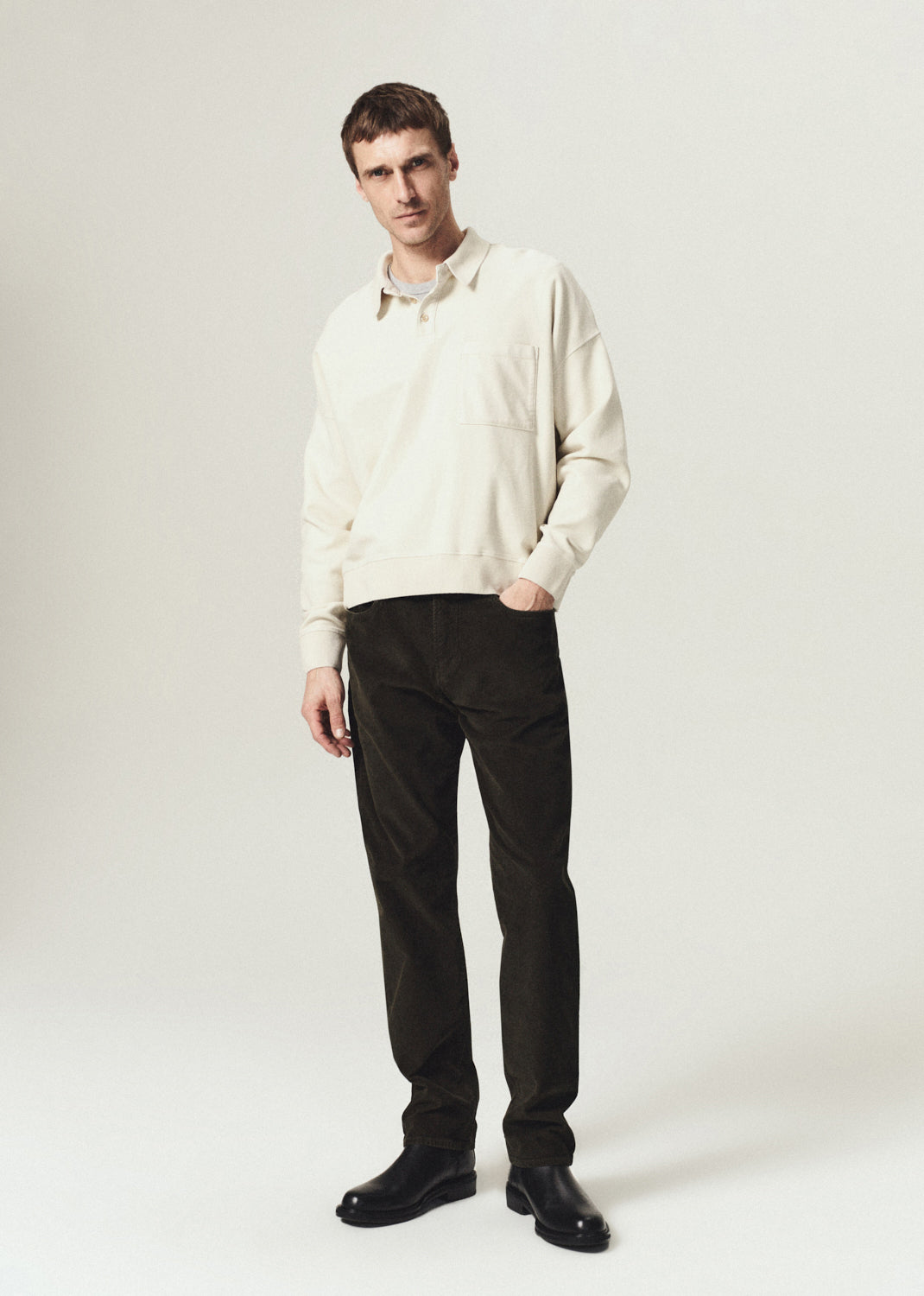 Man wearing an ecru long sleeve polo with dark brown straight leg pants on a grey background.