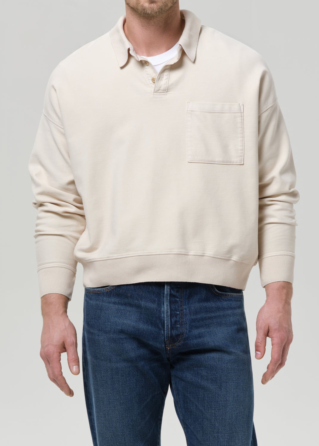 A man wearing a cream-colored long-sleeve rugby-style sweater with a chest pocket, layered over a white t-shirt and paired with blue jeans, shown against a grey background.