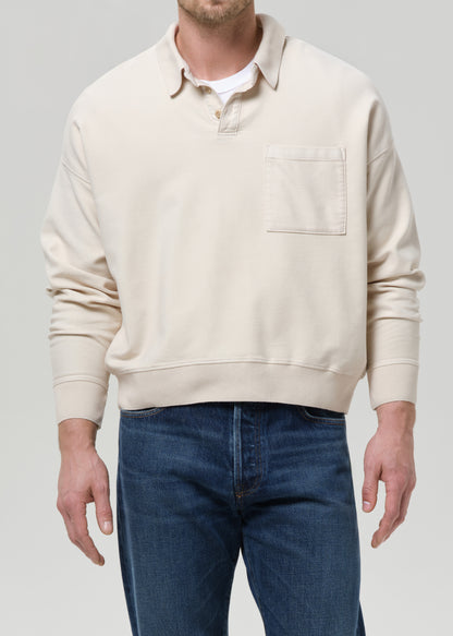 A man wearing a cream-colored long-sleeve rugby-style sweater with a chest pocket, layered over a white t-shirt and paired with blue jeans, shown against a grey background.