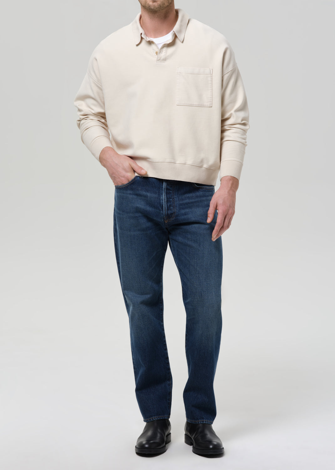A man wearing a cream-colored long-sleeve knit polo sweater, dark blue jeans, and black leather shoes, standing against a grey background.