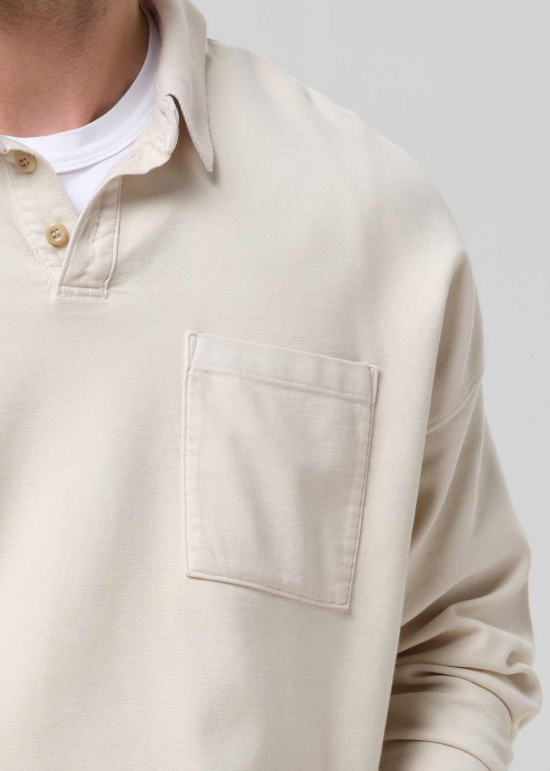 Close-up of a person wearing a light beige long-sleeve polo shirt with a chest pocket over a white undershirt, against a grey background.