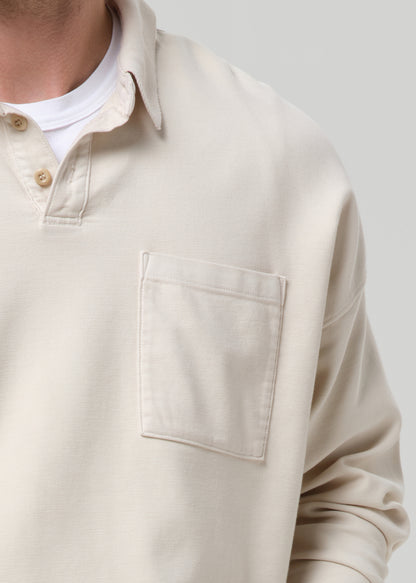Close-up of a person wearing a light beige long-sleeve polo shirt with a chest pocket over a white undershirt, against a grey background.