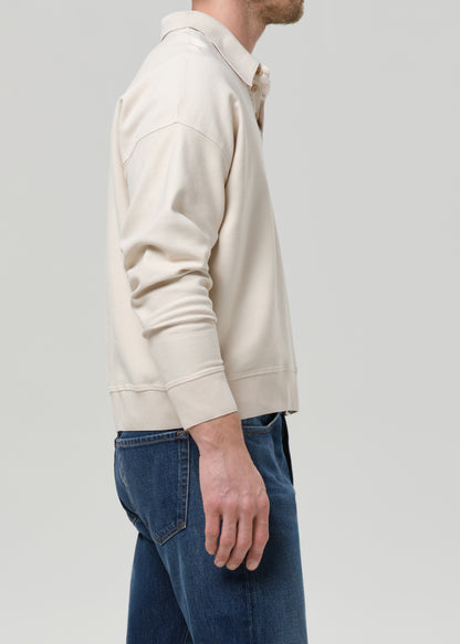 A man shown from the side wearing a cream-colored long-sleeve knit polo-style top and blue jeans, photographed against a grey background.