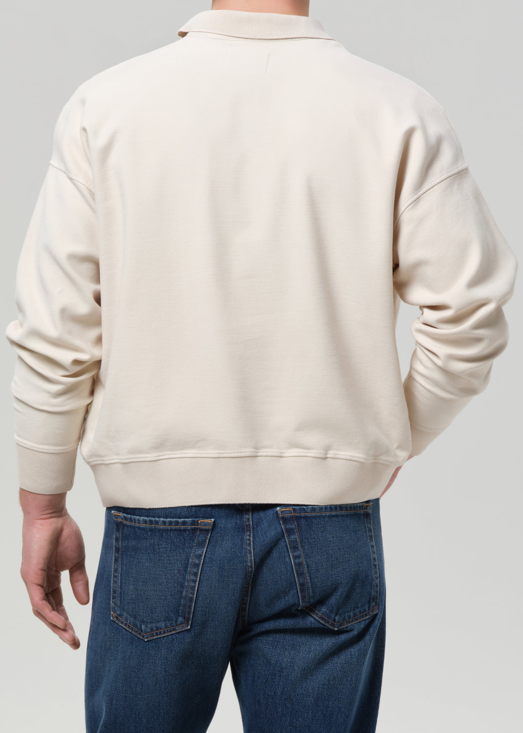 A person shown from the back wearing a plain cream sweatshirt and blue jeans against a grey background.