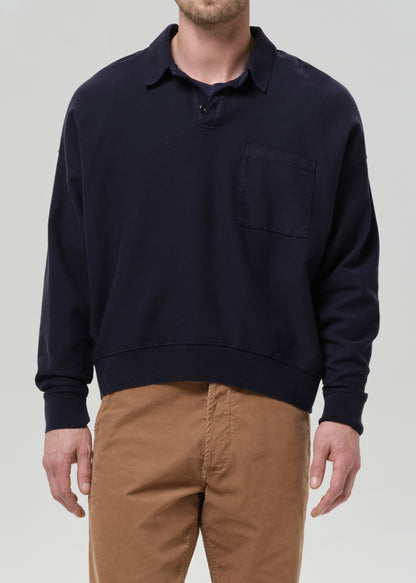A man wearing a dark navy blue long-sleeve knit polo sweater with a chest pocket, paired with brown pants, shown from the waist up against a grey background.