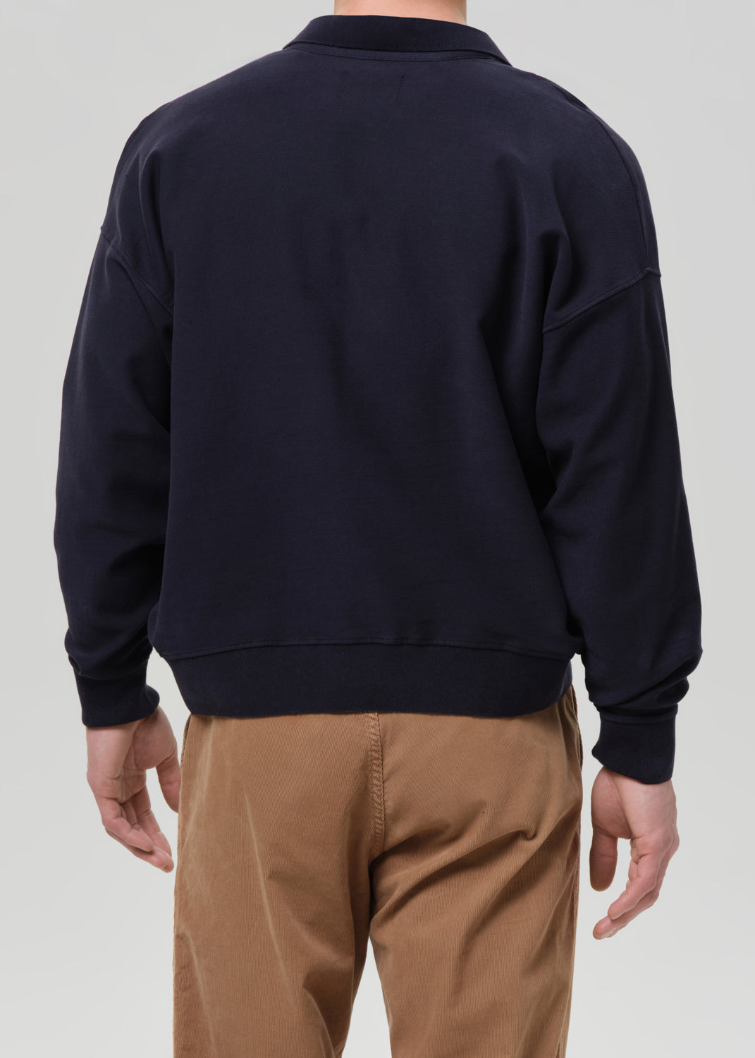 A man viewed from the back wearing a dark navy crewneck sweatshirt and tan pants, set against a grey background.