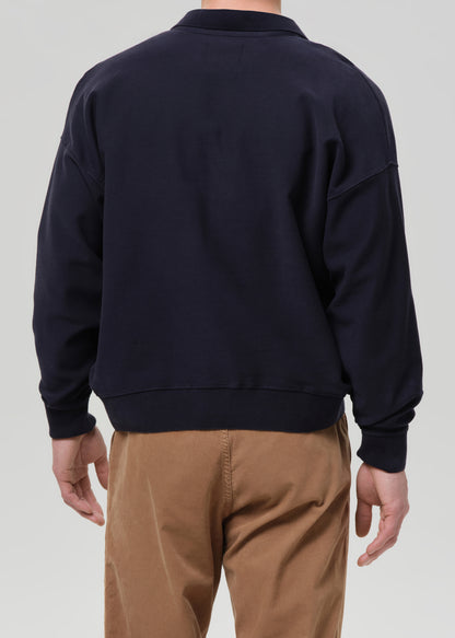 A man viewed from the back wearing a dark navy crewneck sweatshirt and tan pants, set against a grey background.