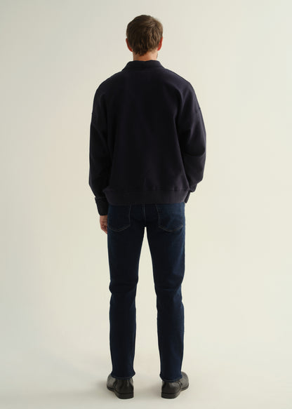 Person wearing a dark navy long sleeve collared pullover with dark blue jeans and black boots, back full body view on a light grey background.