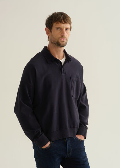 Person wearing a dark navy long sleeve collared pullover with dark blue jeans, angled front cropped view on a light grey background.