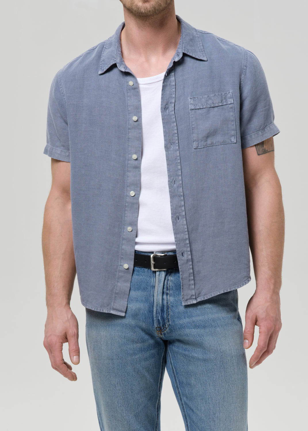 A man wearing a short-sleeve button-up shirt in a dusty blue chambray fabric, layered over a white t-shirt and paired with blue jeans, photographed against a grey background.