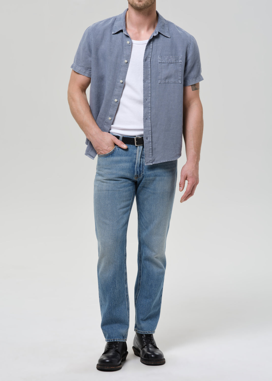 A man modeling a light blue short-sleeve button-up shirt over a white t-shirt, paired with light blue jeans and black leather shoes, against a grey background.