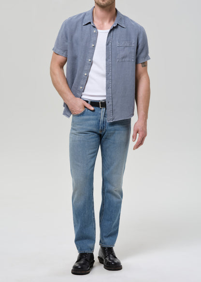 A man modeling a light blue short-sleeve button-up shirt over a white t-shirt, paired with light blue jeans and black leather shoes, against a grey background.