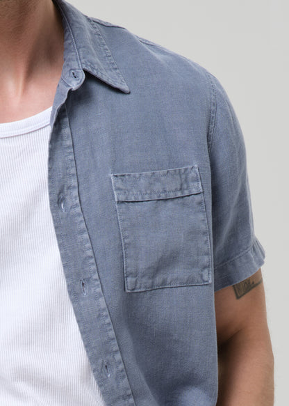 Close-up of a person wearing a short-sleeve button-up shirt in a muted blue-gray color over a white ribbed undershirt, against a grey background.