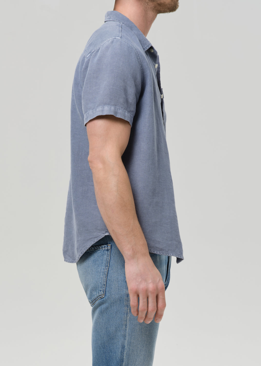 Side profile of a man wearing a short-sleeve button-up shirt in a dusty slate blue color, paired with light-wash blue jeans, against a grey background.