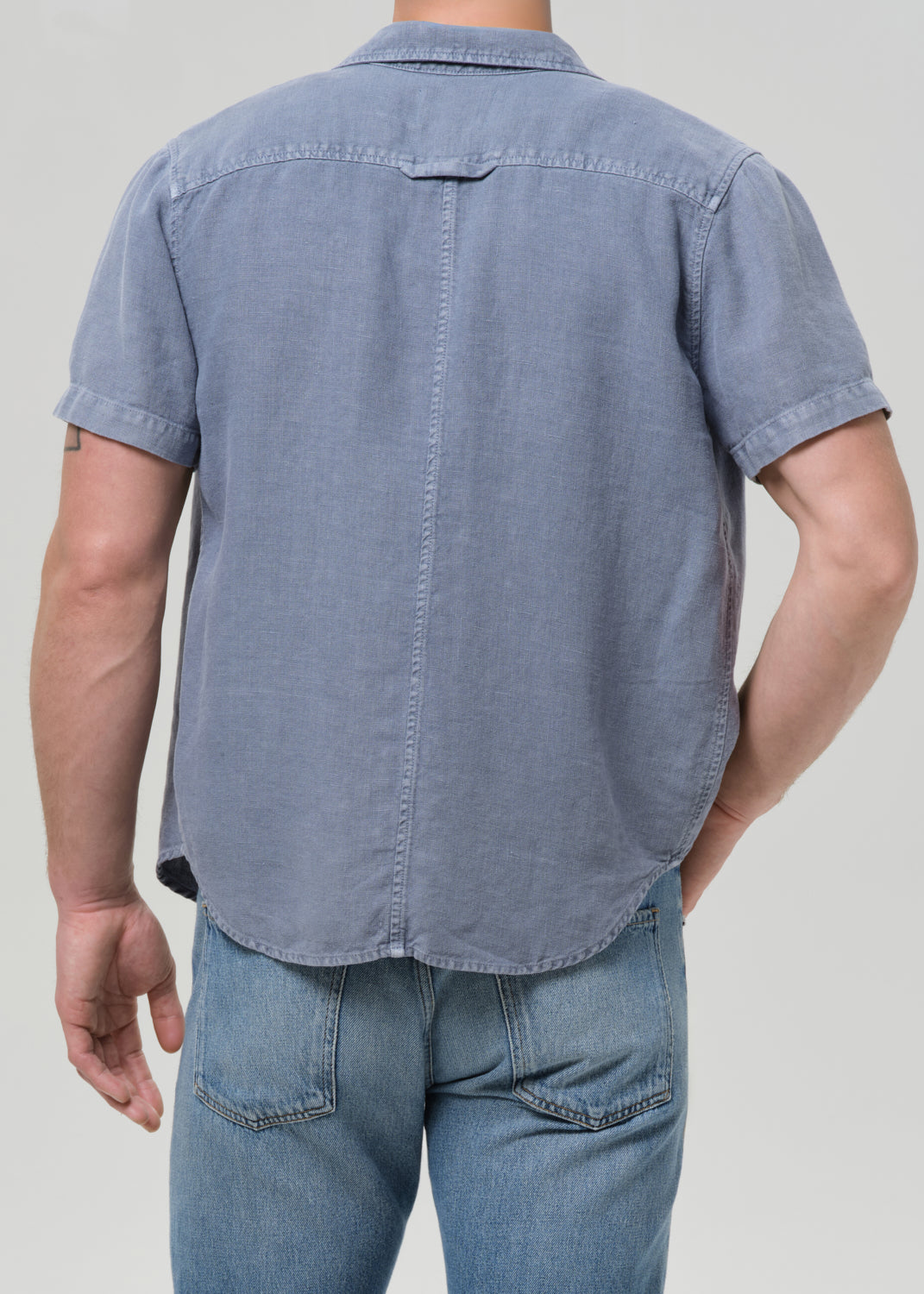 A man wearing a light blue short-sleeve button-up shirt and blue jeans, shown from the back against a grey background.