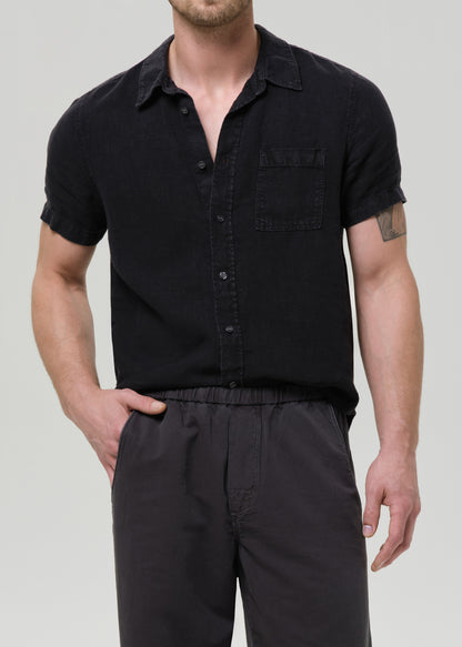A man wearing a black short-sleeve button-up shirt with a chest pocket and dark trousers, photographed against a grey background.
