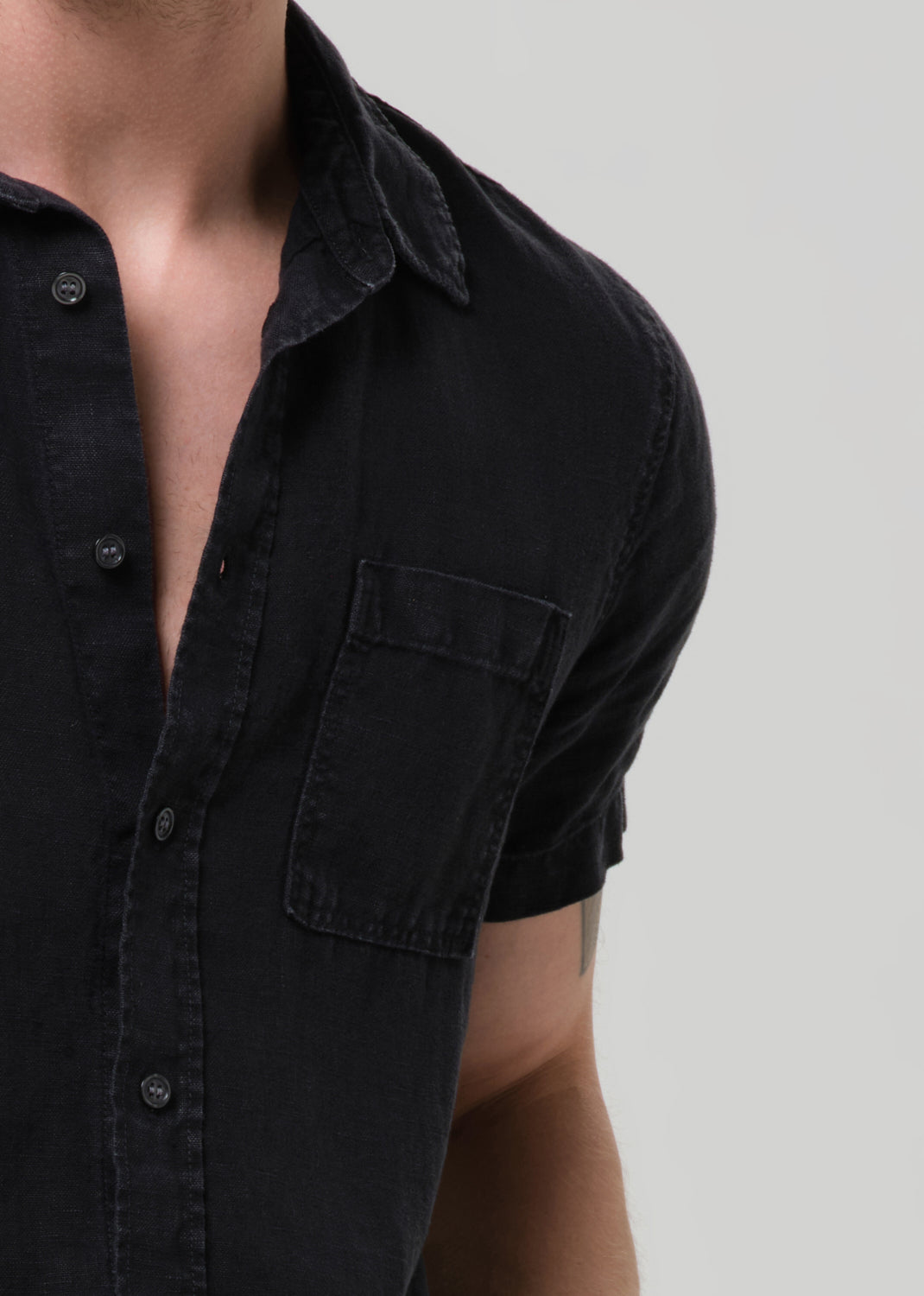Close-up of a person wearing a short-sleeve black button-up shirt with a chest pocket, shown against a grey background.