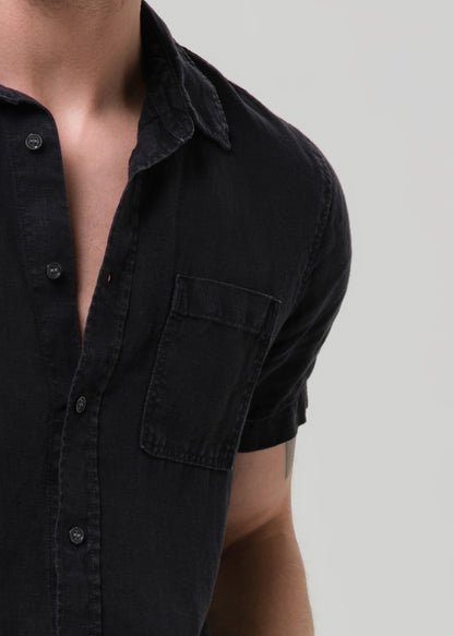 Close-up of a person wearing a short-sleeve black button-up shirt with a chest pocket, shown against a grey background.