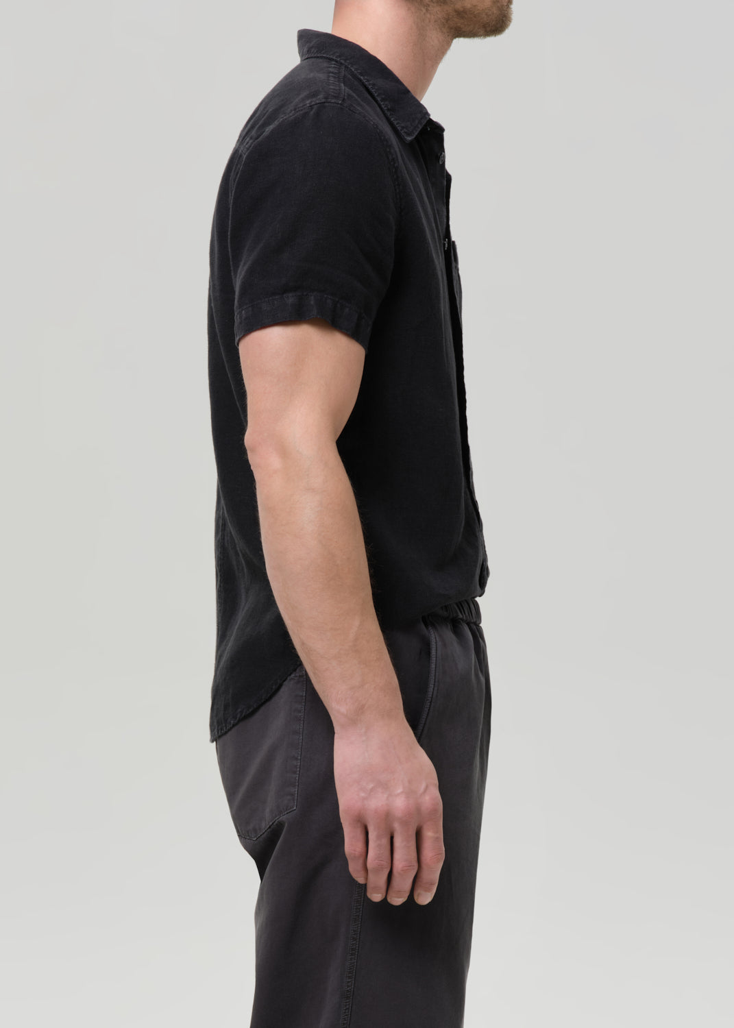 Side profile of a man wearing a black short-sleeve button-up shirt and dark trousers against a grey background.