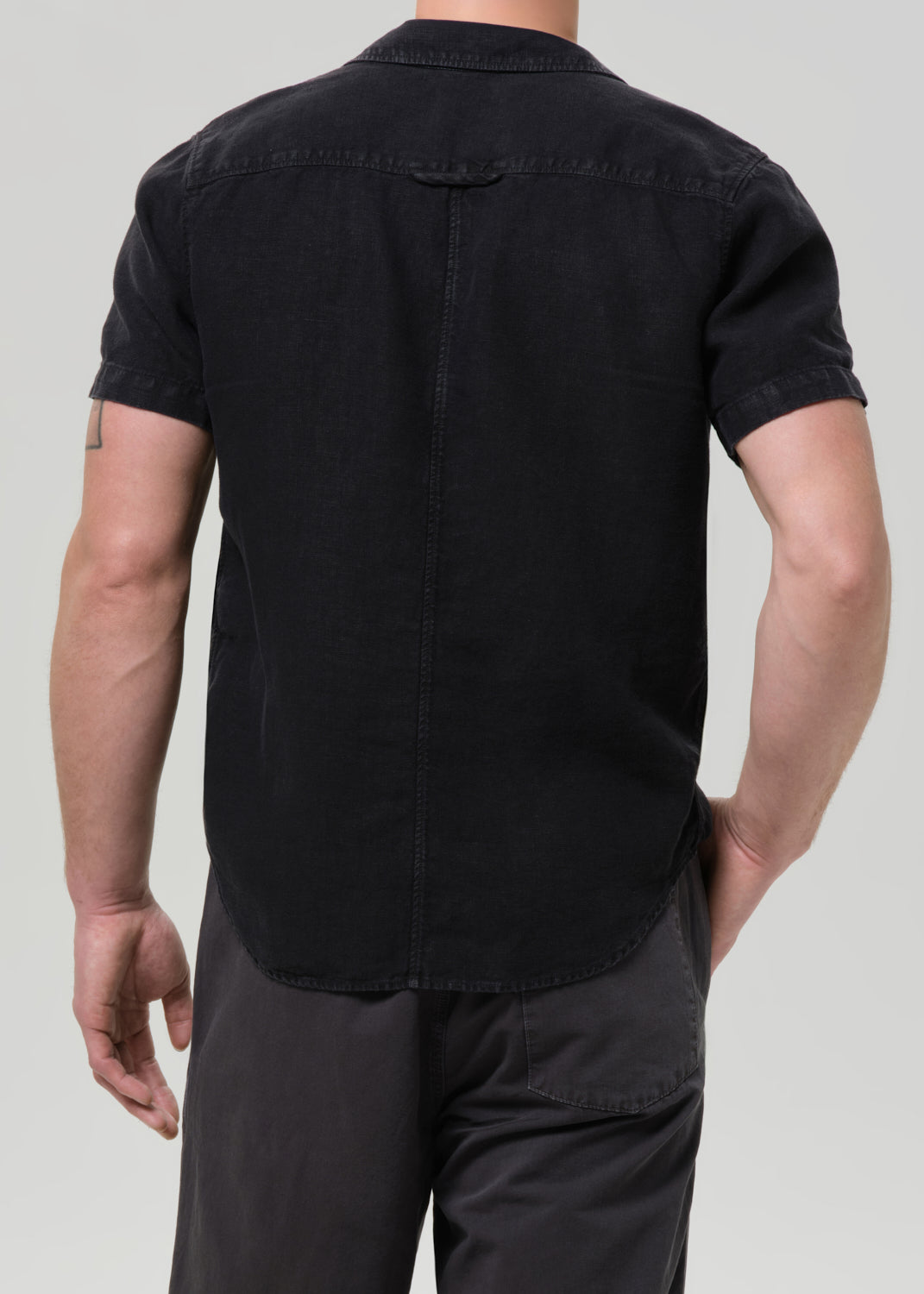 Rear view of a man wearing a black short-sleeve button-up shirt and dark grey pants against a grey background.