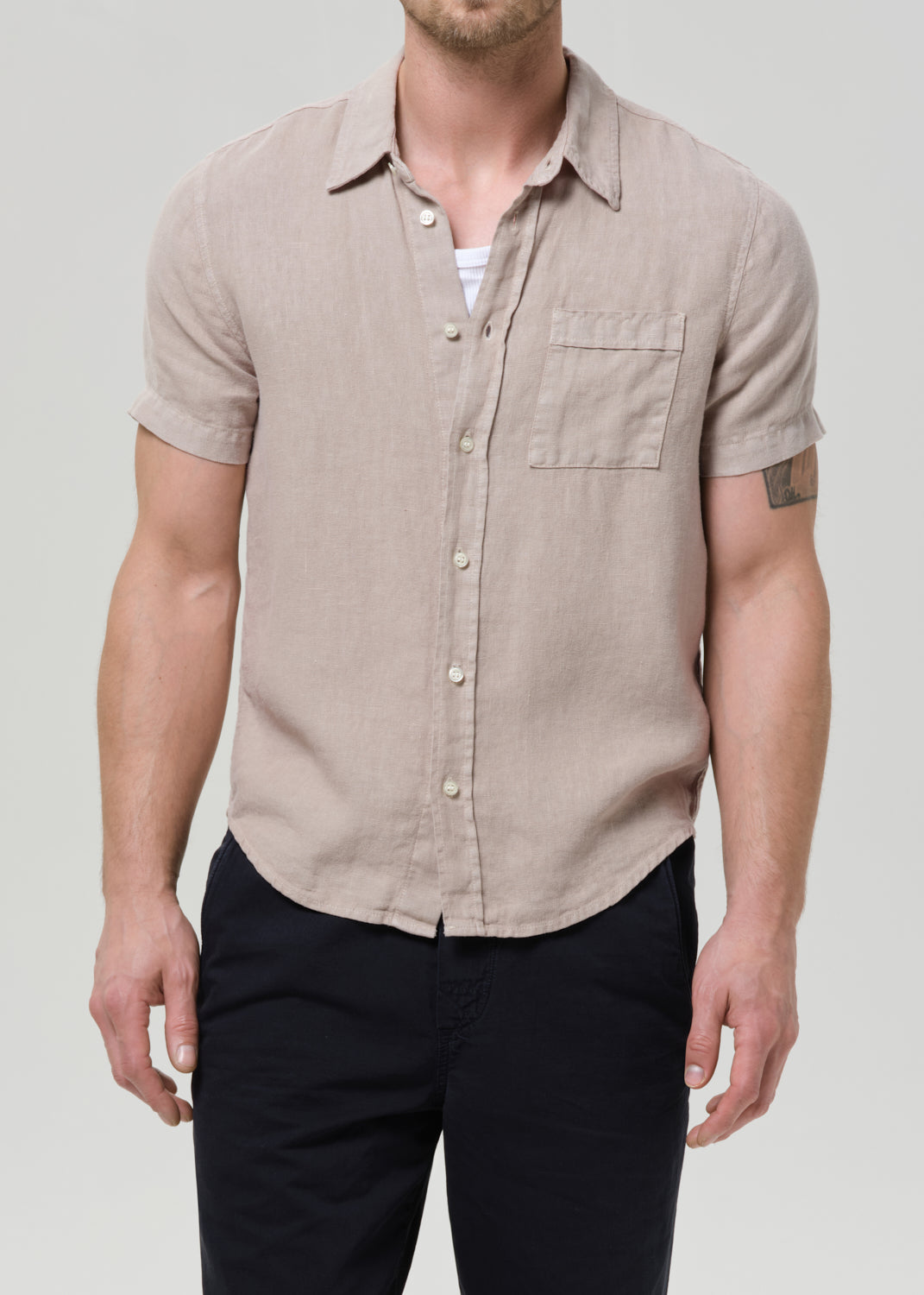 A man wearing a light beige short-sleeve linen button-up shirt with a chest pocket, paired with dark pants, against a grey background.