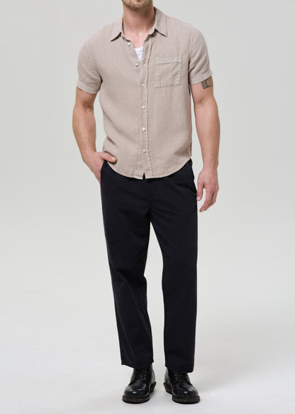 A man modeling a light beige short-sleeve button-up shirt with a chest pocket, paired with black trousers and black shoes, against a grey background.