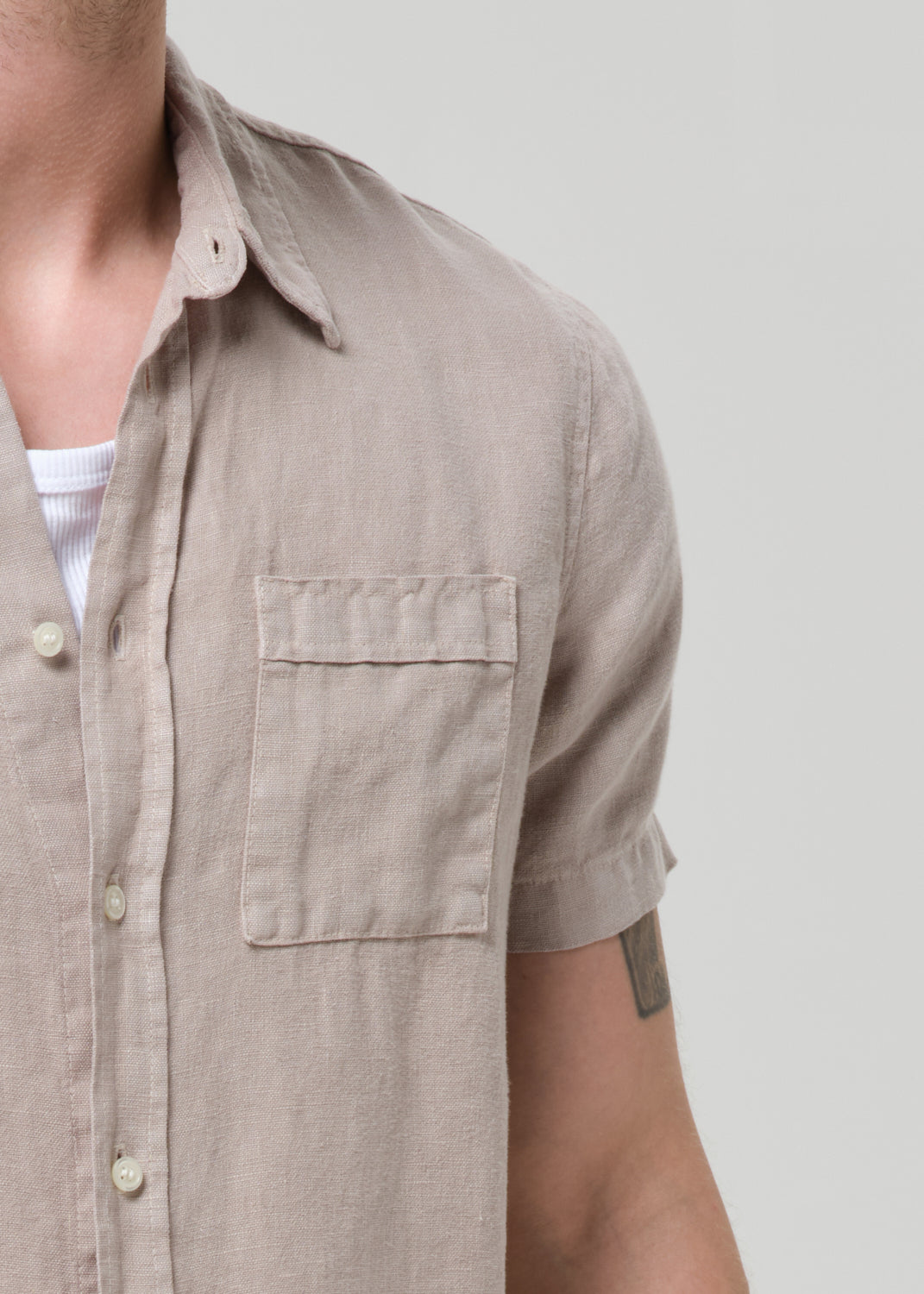 Close-up of a man wearing a light beige short-sleeve linen button-up shirt with a chest pocket, layered over a white undershirt, against a grey background.