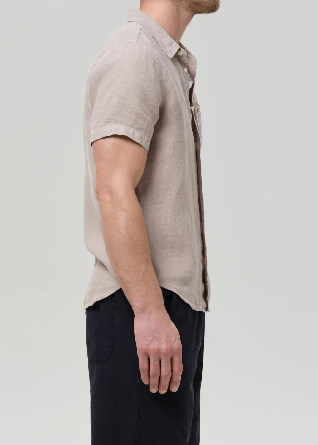 A man shown in profile wearing a light beige short-sleeve linen button-up shirt and dark trousers, photographed against a grey background.
