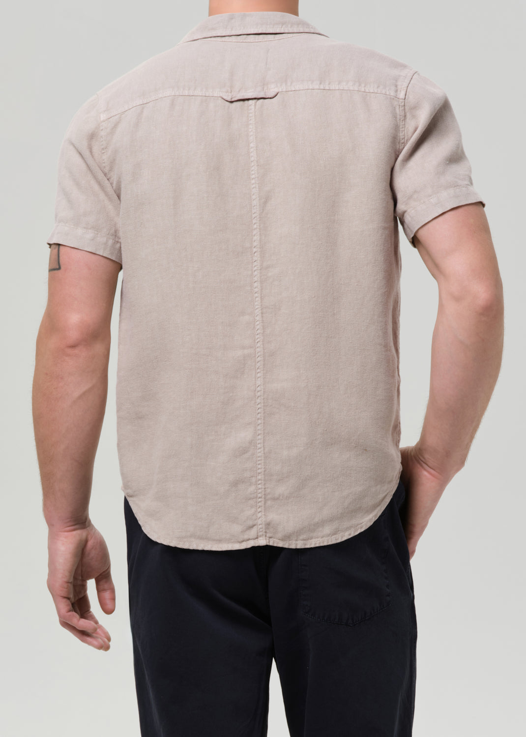 A man viewed from the back wearing a light beige short-sleeve linen button-down shirt and dark navy pants, set against a grey background.