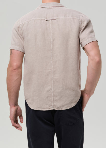 A man viewed from the back wearing a light beige short-sleeve linen button-down shirt and dark navy pants, set against a grey background.