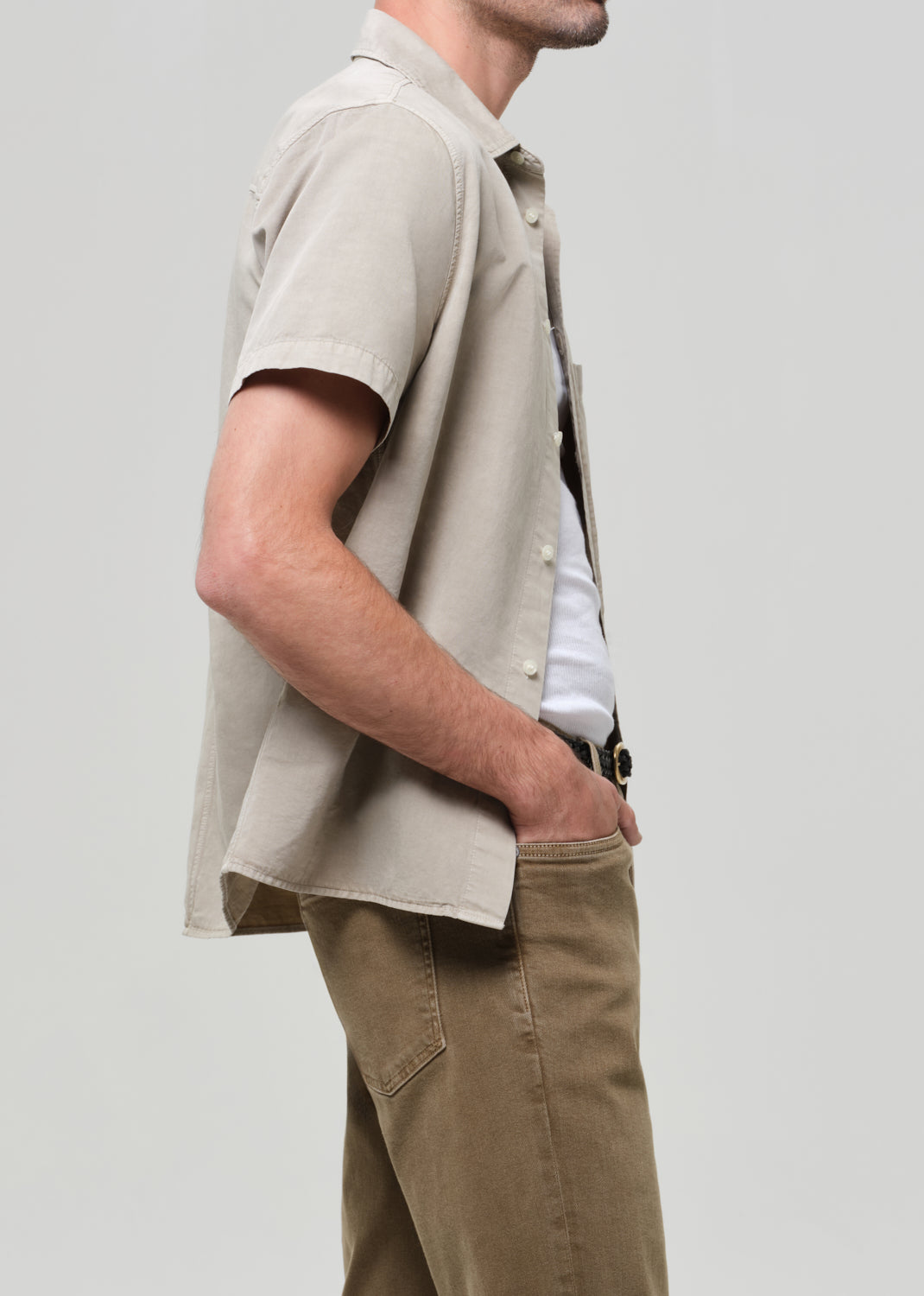 Man wearing a beige short-sleeve shirt over a white t-shirt with brown pants on a light gray background