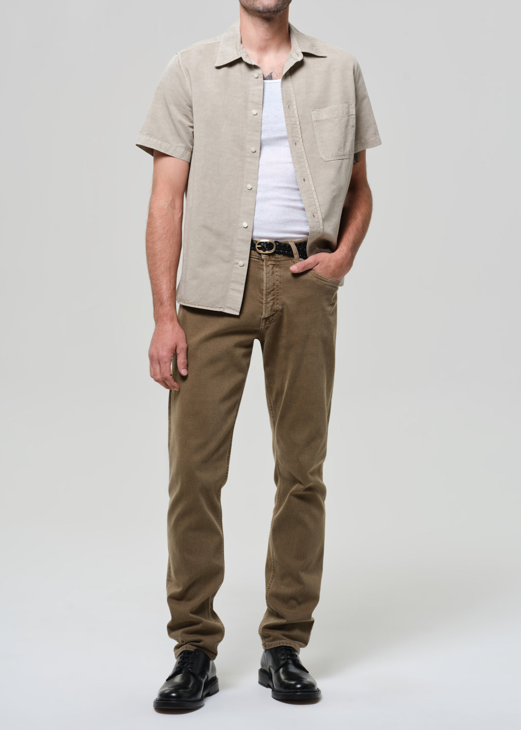Man wearing a beige short-sleeve shirt over a white t-shirt with brown pants on a light gray background