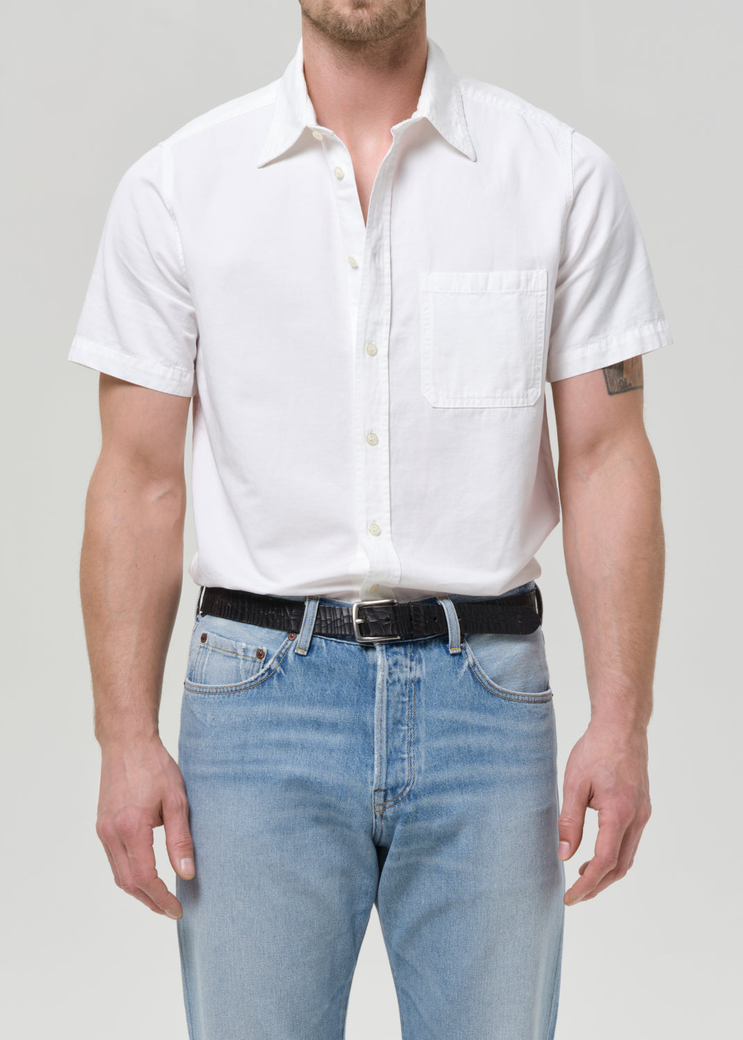 A man wearing a white short-sleeve button-up shirt with a chest pocket, tucked into light blue jeans with a black belt, against a grey background.