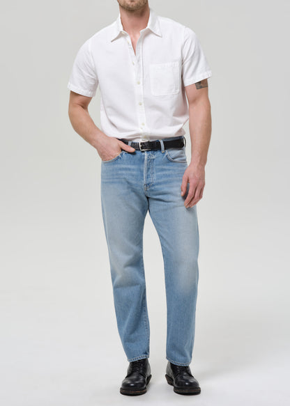 A man modeling a white short-sleeve button-up shirt and light blue jeans with a black belt, standing against a grey background.