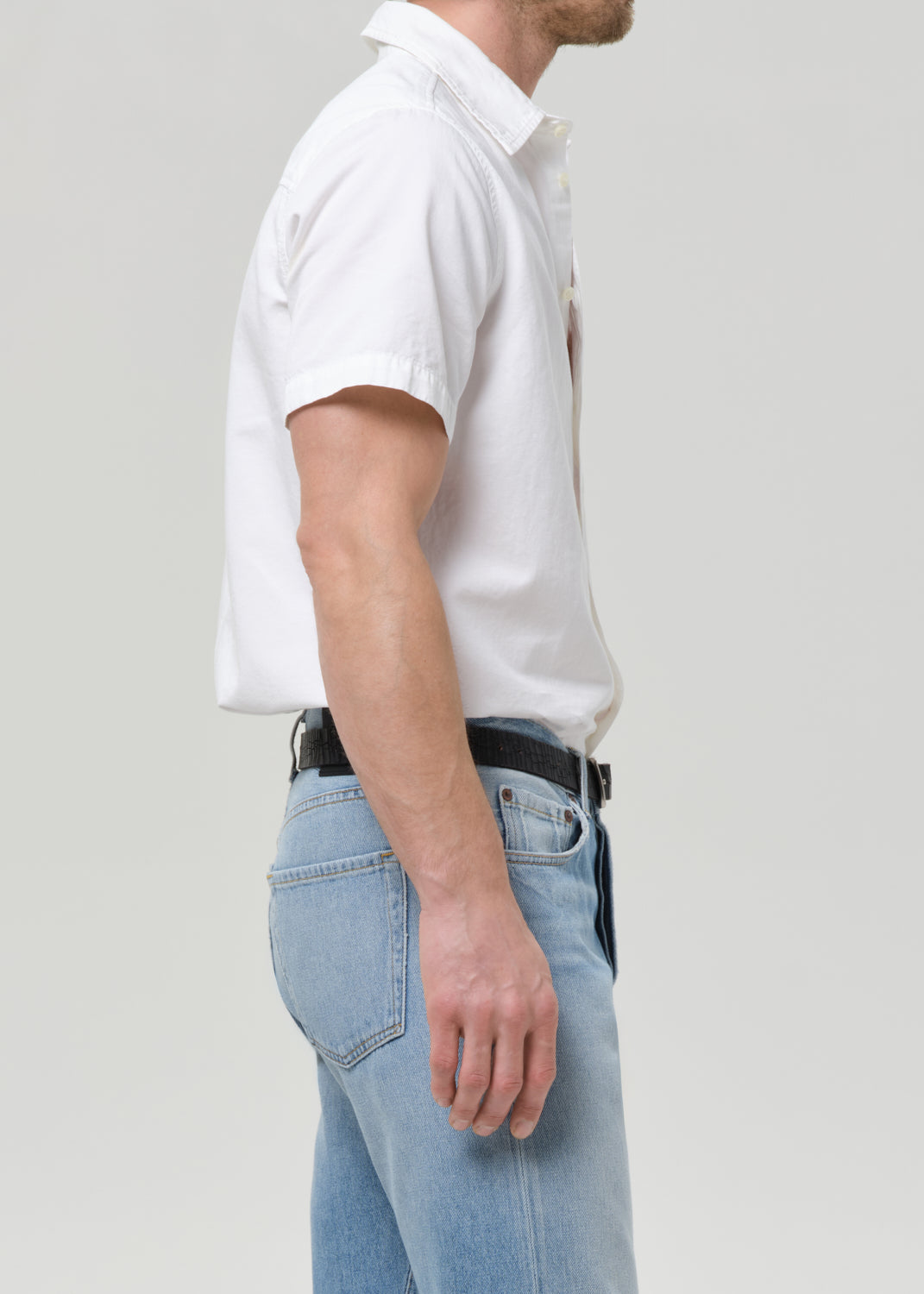 A man shown from the side wearing a white short-sleeve button-up shirt tucked into light blue jeans with a black belt, against a grey background.