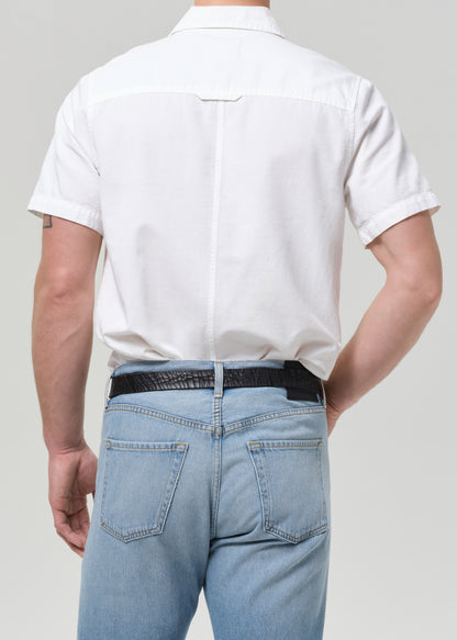 Rear view of a man wearing a white short-sleeve button-down shirt tucked into light blue jeans with a black belt, against a grey background.