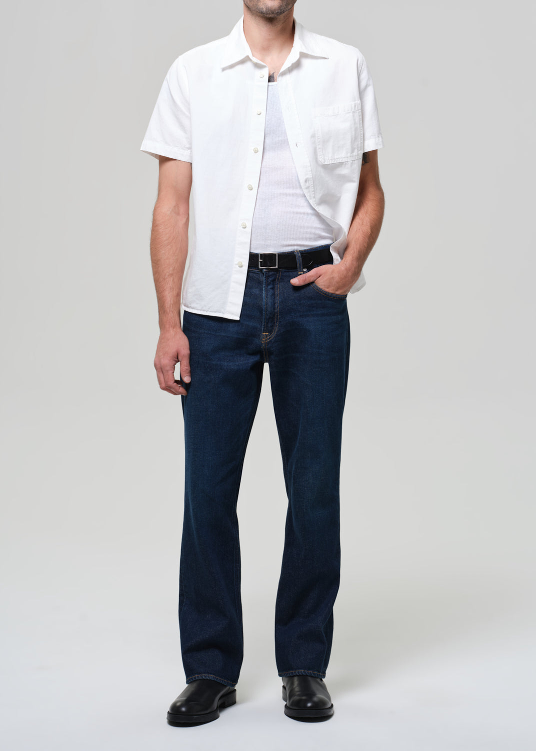 Man wearing a white shirt and dark jeans against a grey background
