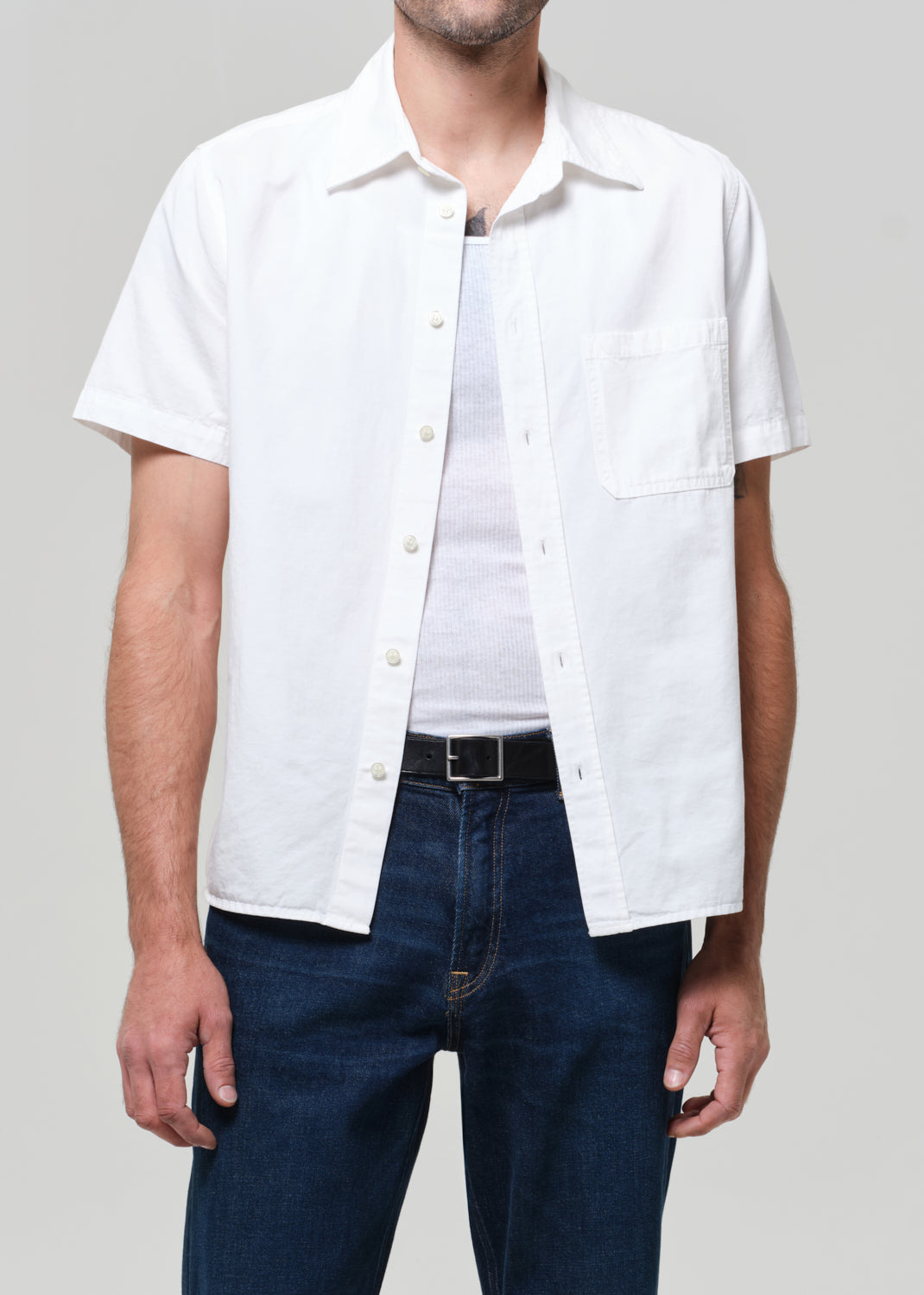 Man wearing a white shirt and dark jeans against a grey background
